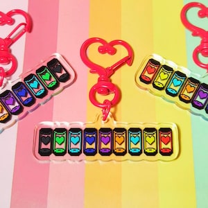 Puede incluir: Llaveros de acrílico transparente con diseños pixelados de corazones coloridos dentro de pequeños recipientes. Cada llavero tiene un cierre rojo en forma de corazón. Los llaveros están dispuestos sobre un fondo degradado de rosa, amarillo y naranja.
