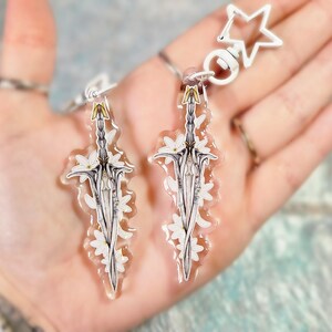May include: Two clear acrylic sword-shaped keychains with white floral accents and gold details. Each keychain has a silver chain and a white star-shaped clasp. The keychains are held in a hand.
