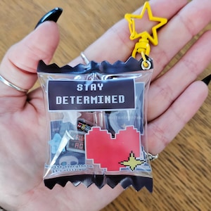 Op de afbeelding: Een doorzichtige plastic zakbedel met een zwarte bovenkant, met diverse kleine bedels en een rood gepixeld hart. De tas heeft de tekst "STAY DETERMINED" en een gele stervormige clip.