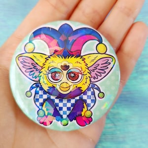 May include: A colourful button featuring a cartoon character with a jester hat, yellow fur, and large blue eyes. The character is wearing a checkered purple and white costume. The button has a holographic finish.