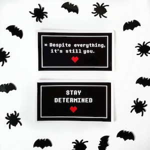 May include: Two black and white stickers with pixelated text. The top sticker says "* Despite everything, it's still you." with a red heart. The bottom sticker says "STAY DETERMINED" with a red heart.