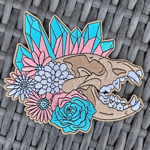 May include: Embroidered patch featuring a tan animal skull with open jaws, adorned with turquoise and pink crystals, and various flowers in shades of pink, white, and turquoise. The patch has a tan border.
