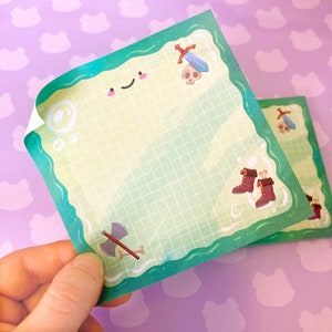 May include: A green and white gridded notepad with a whimsical illustration of a sword, boots, and an axe. The notepad has a green border and a white background.