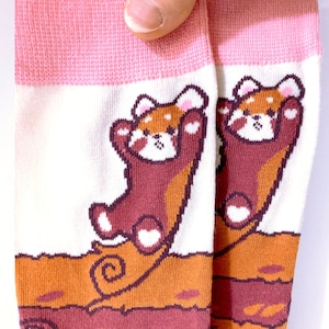 May include: A pair of socks with a repeating pattern of a cartoon red panda with its arms raised. The socks have a white background with a brown and orange design at the bottom and a pink cuff.