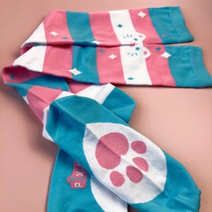 May include: A pair of colourful socks with a pink, white, and teal striped pattern. The socks feature a paw print design on the foot and a small graphic near the ankle. The socks are made of a soft, stretchy material.