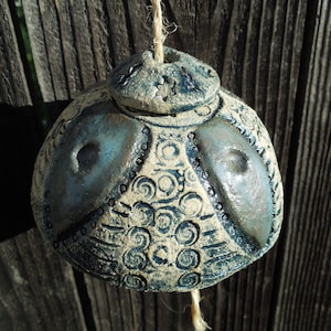 May include: A round, textured ceramic bell with a blue and beige finish, featuring intricate stamped spiral and dot patterns. The bell has a small, separate ceramic cap at the top and is suspended by a thick, light-colored natural fiber twine.