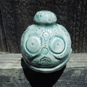 May include: A ceramic figurine with a blue-green glaze and intricate patterns. The figurine has a round body with two large eyes and a small mouth.