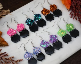 May include: Five pairs of dangle earrings featuring black cauldron charms topped with glittery resin bubbles in orange, teal, pink, purple, and lime green. Each earring hangs from a silver-toned metal hook, showcasing a festive design.
