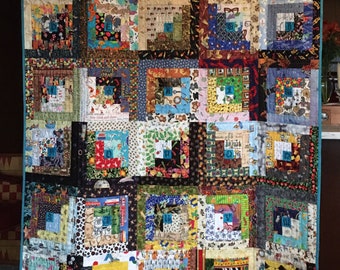 May include: A colorful quilt with a log cabin pattern. The quilt is made up of squares of different fabrics, each with a different pattern. The quilt is bordered with a green and blue trim.