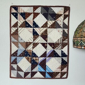 May include: A small, square quilt with a patchwork design. The quilt features a variety of fabrics in shades of brown, blue, and white, arranged in a geometric pattern. The quilt is bordered with a dark brown fabric and has a quilted texture.