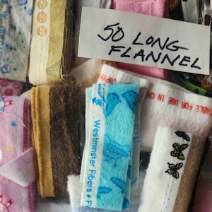 May include: A collection of colorful fabric swatches, including pink, blue, brown, and white. A handwritten sign reads "50 LONG FLANNEL". Some swatches have printed patterns and text, such as "Westminster Fibers # F".