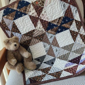 May include: A handmade patchwork quilt with a variety of patterned fabrics in shades of brown, white, and blue. The quilt is adorned with intricate quilting patterns and a brown border. A small teddy bear sits next to the quilt.