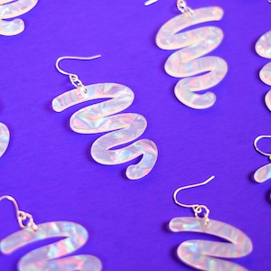 Puede incluir: Un par de pendientes blancos iridiscentes con forma de espiral. Los pendientes están hechos de un material plástico transparente con un acabado nacarado. Los pendientes cuelgan de ganchos plateados.