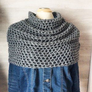 May include: A gray crocheted cowl neck warmer. The handmade neck warmer is displayed on a mannequin and worn over a denim jacket. The cowl is a solid gray color and has a textured, open weave design.
