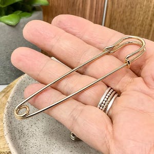以下が含まれることがあります： 大きな金色の安全ピン。ピンは手で持たれており、留め金と先端がはっきりと見えます。ピンは光沢のある金属でできており、デザインはシンプルでエレガントです。ピンは装飾的なアクセサリーです。