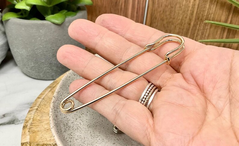 以下が含まれることがあります： 大きな金色の安全ピン。ピンは手で持たれており、留め金と先端がはっきりと見えます。ピンは光沢のある金属でできており、デザインはシンプルでエレガントです。ピンは装飾的なアクセサリーです。