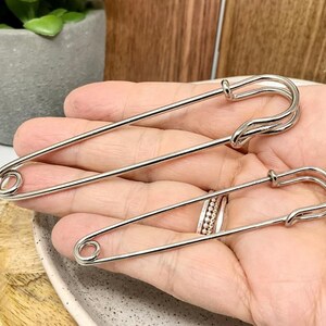 以下が含まれることがあります： サイズが異なる2つの銀色の安全ピン。 シンプルでありながら機能的なデザインが、手で示されています。 ピンは、安全な留め金が付いた、クラシックな湾曲した形状をしています。
