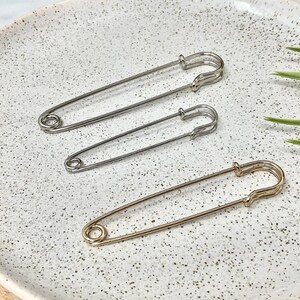 以下が含まれることがあります： シルバーとゴールドの3つの安全ピン。ピンは、背景に緑の植物の葉がいくつかある、斑点のあるオフホワイトの表面に配置されています。ピンのサイズは異なります。裁縫やクラフトに役立つアイテムです。