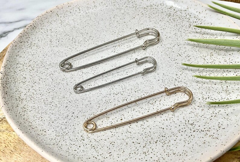 以下が含まれることがあります： シルバーとゴールドの3つの安全ピン。ピンは、背景に緑の植物の葉がいくつかある、斑点のあるオフホワイトの表面に配置されています。ピンのサイズは異なります。裁縫やクラフトに役立つアイテムです。
