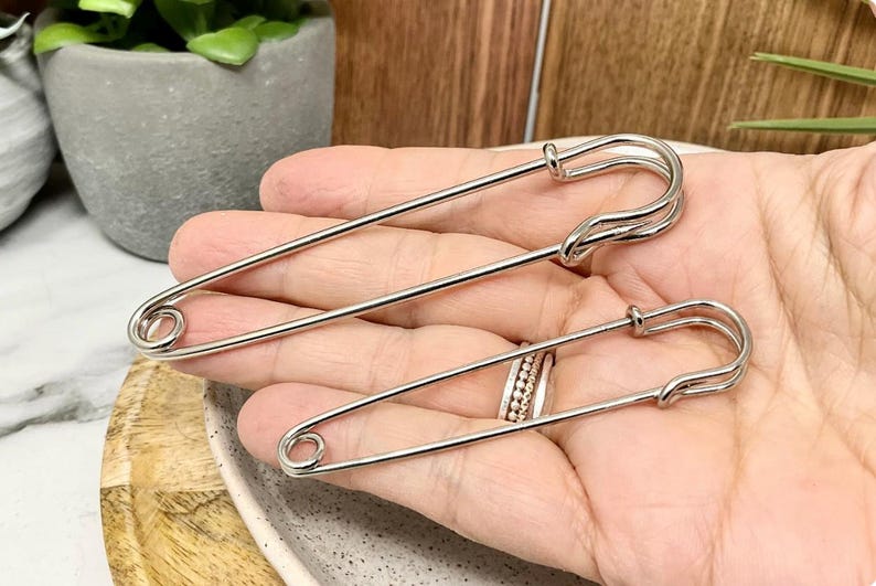 以下が含まれることがあります： サイズが異なる2つの銀色の安全ピン。 シンプルでありながら機能的なデザインが、手で示されています。 大きなピンは約10cm、小さなピンは約5cmです。