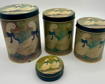 May include: Set of four round metal canisters with a green and brown background featuring a winter scene with white ducks wearing blue bows. The canisters are different sizes and have a gold trim around the top.