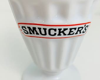 May include: White ceramic Smucker's branded milkshake glass with a fluted design. The glass has a wide base and a narrow top.