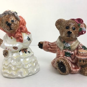 Puede incluir: Un par de figuritas de osos de peluche de cerámica. La osa lleva un vestido blanco con un lazo rojo y un sombrero. El oso lleva una camisa a rayas marrones y rosas y sostiene una taza de té.