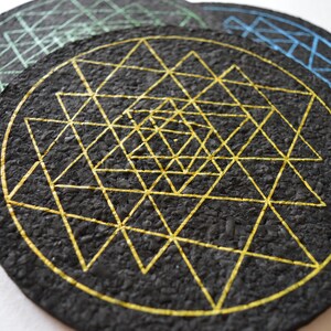 May include: Three round cork coasters with geometric designs. The top coaster has a black background with a gold geometric design. The middle coaster has a green geometric design on a black background. The bottom coaster has a blue geometric design on a black background.