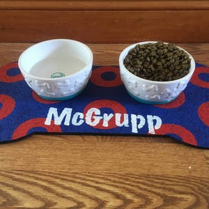 May include: A blue and red bone-shaped dog food mat with the name "McGrupp" printed in white. Two white ceramic dog bowls are on the mat, one filled with dog food.