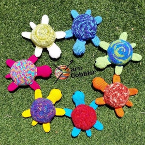 Puede incluir: Siete juguetes de tortuga de crochet en varios colores, incluyendo azul, verde, rojo, amarillo y blanco. Las tortugas están dispuestas en círculo sobre una superficie de hierba verde. El texto "Yarn Gobbler" es visible en el centro de la imagen.