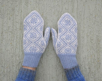 May include: A pair of light blue and white knitted mittens. The mittens feature a geometric diamond pattern with a snowflake design. The cuffs and the base of the mittens are a solid light blue color. The mittens are displayed against a textured gray background.