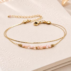 Puede incluir: Una pulsera dorada de doble cadena que presenta una hilera de pequeñas cuentas facetadas de color rosa pálido intercaladas con diminutos espaciadores metálicos dorados. La pulsera incluye un cierre de mosquetón y una cadena extensora.