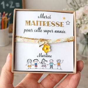Könnte beinhalten: Ein Armband aus einer goldfarbenen Kette und Stoffband mit einem Herz- und einem gelben Blumenanhänger, präsentiert auf einer weißen Karte mit dem Text "Merci MAÎTRESSE pour cette super année." und "Marlène" über einer Zeichnung von fünf Kindern.