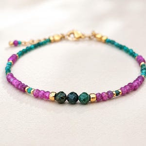 Op de afbeelding: Een delicate kralenarmband met afwisselende segmenten van gefacetteerde magenta en blauwgroene edelsteenkralen. Het ontwerp bevat kleine goudkleurige metalen tussenkralen en een centraal middelpunt van drie grotere, donkergroene gefacetteerde kralen.