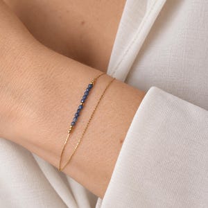 Op de afbeelding: Een verfijnde dubbele gouden kettingarmband met een rij kleine, gefacetteerde blauwe edelsteenkralen, geaccentueerd door piepkleine gouden bolletjes. De armband bestaat uit een effen gouden ketting en een ketting versierd met blauwe kralen.