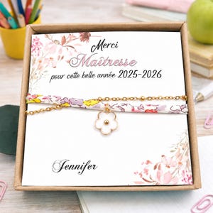 Könnte beinhalten: Ein goldfarbenes Kettenarmband mit einem floralen Stoffband und einem weißen Blumenanhänger mit goldener Mitte, präsentiert auf einer weißen Karte mit Blumenillustrationen und dem Text Merci Maîtresse pour cette belle année 2025-2026 und Jennifer.
