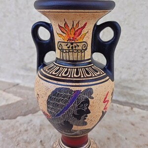 Pode incluir: Uma pequena ânfora de cerâmica preta e castanho com duas alças. A ânfora é decorada com uma silhueta preta de uma figura com uma faixa azul, uma chama vermelha e amarela e um padrão geométrico preto e branco.