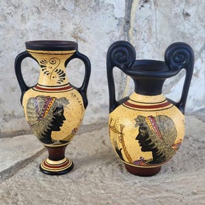 Pode incluir: Dois vasos gregos de cerâmica preta e amarela com figuras pintadas e detalhes vermelhos. O vaso menor tem duas alças e o maior tem duas alças com pontas em espiral.