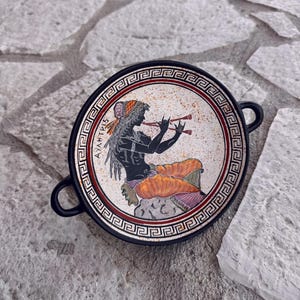 May include: A black and white ceramic plate with a red and white border. The plate features a black and white illustration of a woman playing a double flute. The text "AYANTIS" is written on the plate.