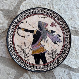 Könnte beinhalten: Ein runder dekorativer Keramikteller mit der schwarzen Silhouette einer Figur, die Pfeil und Bogen hält, beschriftet mit APTEMIS. Die Figur trägt einen gelben Rock und eine violette Schärpe, begleitet von einem kleinen Vogel und stilisierter Flora.