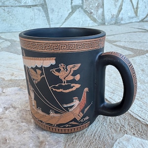 Puede incluir: Una taza de cerámica negra mate con una ilustración en color terracota de un barco antiguo con remos y vela. Sobre el barco, una sirena alada toca la flauta. El borde y el asa están decorados con un patrón geométrico de greca griega.
