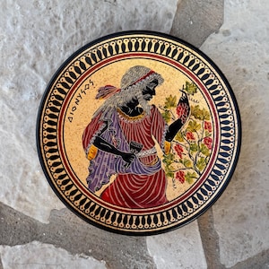 May include: A round, decorative plate featuring a classical Greek illustration. The central image depicts a figure in red and purple robes holding grapes. The plate has a gold background and a black border with a repeating pattern.