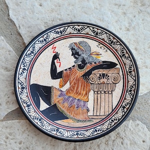 Puede incluir: Un plato de cerámica blanco y negro con un diseño rojo, naranja y morado de un dios griego sosteniendo un rayo. El plato tiene un borde negro con un borde rojo y blanco. El texto "ZEUS" está escrito en blanco en el plato.