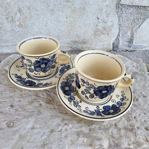 May include: Two ceramic teacups with blue floral designs and matching saucers. The cups and saucers have a cream-colored background.
