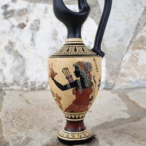 May include: A decorative ceramic jug with a black handle and spout. The jug features a beige body with a painted illustration of a figure in red and black, with the word "ARTEMIS" visible. The base is dark red and the jug is on a stone surface.