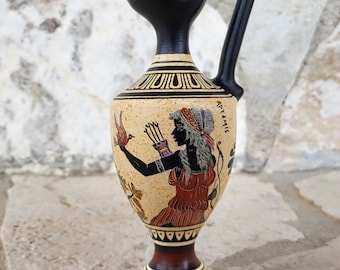 Op de afbeelding: Een decoratieve keramische kruik met een zwarte handgreep en tuit. De kruik heeft een beige lichaam met een geschilderde illustratie van een figuur in rood en zwart, met het woord "ARTEMIS" zichtbaar. De basis is donkerrood en de kruik staat op een stenen oppervlak.
