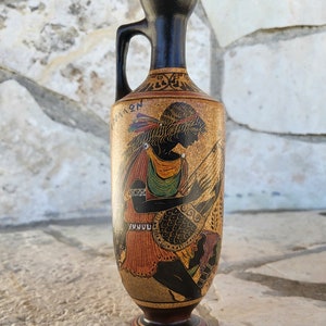 Può includere: Un vaso in ceramica nero e marrone con un manico. Il vaso è decorato con un'immagine in bianco e nero di una donna che suona una lira. Il vaso ha la scrittura greca.