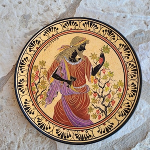 Può includere: Un piatto rotondo in ceramica con un bordo nero e uno sfondo dorato. Il piatto presenta un'illustrazione in bianco e nero di una figura che tiene dell'uva, con il testo "DIONYSOS" scritto in nero. Il piatto è decorato con un motivo geometrico nero e dorato.