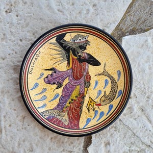 Puede incluir: Un plato redondo de cerámica con un borde negro y un fondo amarillo. El plato presenta una imagen pintada de una figura con una corona, sosteniendo un tridente y montando un caballito de mar. La figura está rodeada de olas azules y amarillas. El texto "ΠΟΣΕΙΔΩΝ" está escrito en griego en el plato.