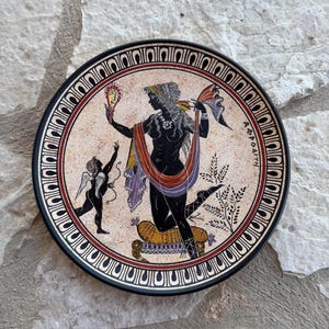 Puede incluir: Un plato decorativo redondo con una ilustración de estilo de figuras negras que muestra a una figura sosteniendo un espejo y un querubín alado. La escena incluye un cojín amarillo, una planta y el texto griego AΦPOΔITH. El borde tiene un patrón.